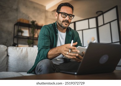 Adult man wear eyeglasses and a green shirt work from home in a modern living room, hold a notepad while use a laptop, suggesting remote work and creativity - Powered by Shutterstock