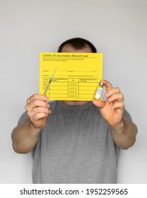 Adult Man Wearing T-shirt Holding Yellow Vaccination Record Card, Syringes, And Vials With COVID-19 Vaccine For Two-stage Injection. Gray Background