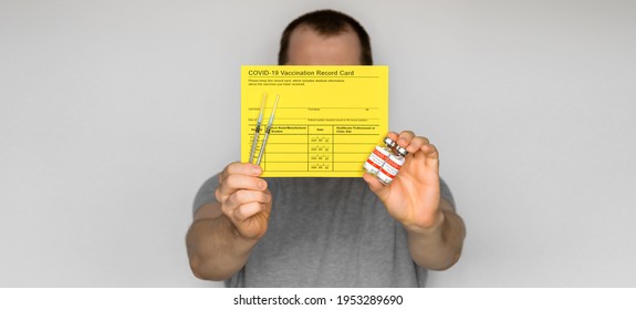 Adult Man Wearing T-shirt Holding Yellow Vaccination Record Card, Syringes, And Vials With COVID-19 Vaccine For Two-stage Injection. Gray Background