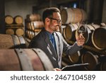 Adult man winemaker hold glass and drink wine stand between the barrels in the cellar check process of make white wine