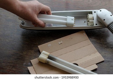 Adult Man's Hand Installs A New Compact Fluorescent Lamp In The Light Fixture. A Gray Table Lamp Rests On A Brown Wooden Table. Replacing The Failed Light Bulb With A New One.