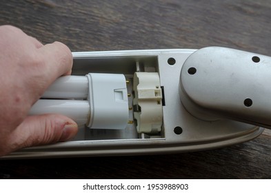 Adult Man's Hand Installs A New Compact Fluorescent Lamp In The Light Fixture. A Gray Table Lamp Rests On A Brown Wooden Table. Replacing The Failed Light Bulb With A New One.