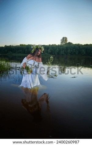 Adult mature brunette woman in a white dress, sundress and a wreath of flowers in summer in water of river or lake in the evening at sunset. Celebration of the Slavic pagan holiday of Ivan Kupala