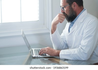 Adult mature Caucasian male doctor in white medical uniform sit at desk in office consult client on laptop online, man GP or physician fill patient medical card use Web application form on computer