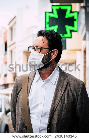 Adult mature caucasian man walk outdoor in the city using healthy protection mask for virus contagion and pharmacy in background - coronavirus new lifestyle concept