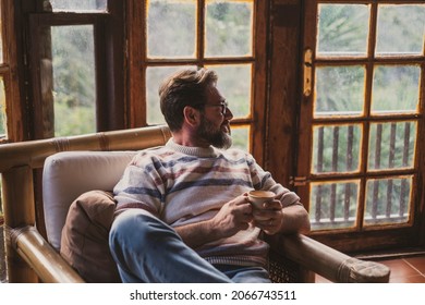 Adult mature man sitting on the chair drinking tea and looking outside the windows at home. Concept of people in indoor leisure activity and relax time. Bearded hipster male alone - Powered by Shutterstock