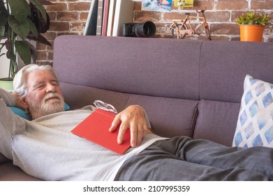Adult mature white haired senior man lying on sofa with closed eyes at home with hand on a book. Attractive bearded retired grandfather enjoying free time and retirement, leisure and relax concept