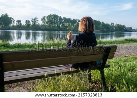 adult mature Woman smoking with her legs crossed sitting on a park bench near a lake in springtime