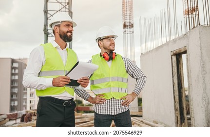 Adult men in waistcoats and hardhats smiling and looking away while working on construction site in city together