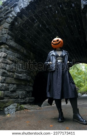 An adult model wearing a pumpkin head mask poses for a full body portrait with hands in their pockets in front of the Glen Span Arch in Central Park
