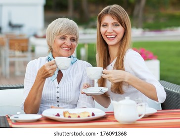 Adult Mother And Daughter Drinking Tea Or Coffee. They Communicate Sitting Outside In A Cafe.