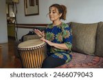 An Adult Percussionist Latin Woman at home studying music with a djembe drum. she have an African style shirt, The scene is relaxed and peaceful, with the woman enjoying her music moving her hands.