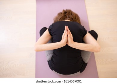 Adult Person Does Yoga Asana, Meditation. Sitting On The Mat. Top View. Woman In Sports Uniform Is Doing Yoga Exercises. No Face Visible. Bend Forward. Namaste From Behind. Copy Space. Wooden Floor. 