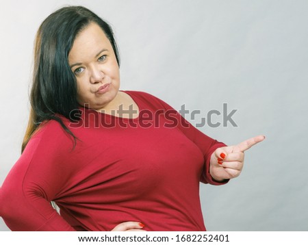 Adult plus size woman pointing with finger at copy space with funny face expression, on grey