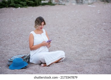 An adult, pregnant woman in a white sundress sits on the sand by the sea, talking on a smartphone. Traveler with backpack, hat. Lifestyle pregnancy tourism concept. Woman sitting in lotus position
