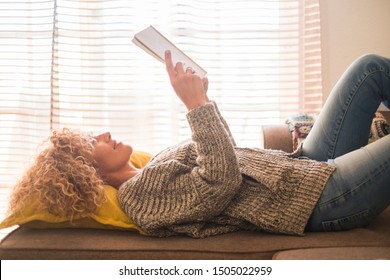 Adult Relaxed Beautiful Curly Blonde Hair Caucasian Woman Lay Down And Read A Paper Book Alone At Home With Window Light In Background - Concept Of Lifestyle And No Stress