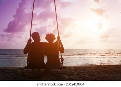 Adult Retired Couple Sitting On Rope Swing And Looking On Sunset Sea Beach. Man Embrace His Wife. Mature Family Relax On Vacation.