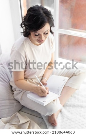 An adult satisfied woman with false eyelashes sits on a wide windowsill. Makes entries in his diary.