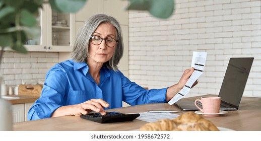 Adult senior 60s woman calculating paper documents at home at pc. Serious middle aged woman at table working on bank payments household online with computer technologies.