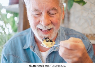 Adult Senior Bearded Caucasian Man Sitting In Cafe Eating A Piece Of Fruitcake With Cream And Blueberries. Happy Elderly Grandfather Enjoying Sweet Food Opens His Mouth Holding A Teaspoon In His Hand