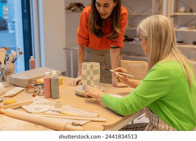 An adult and senior woman focused on painting a checkered pattern on a ceramic vase in a workshop. - Powered by Shutterstock