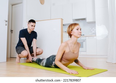 Adult Siblings Doing Yoga At Home. Sister Doing Upward Facing Dog, Brother Holding Her Legs.