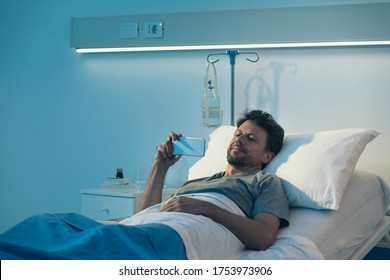 Adult Sleepless Patient Lying In A Hospital Bed At Night, He Is Watching Movies With His Smartphone