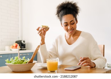 Adult Smiling African Woman Sitting By Table Looking On Her Phone While Eating Avocado Toast And Salad With Glass Of Orange Juice In Cozy Kitchen At Home