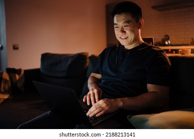 Adult Smiling Asian Man Sitting On Sofa With Laptop And Working In Dark Room At Home