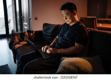 Adult Smiling Asian Man Sitting On Sofa With Laptop And Working In Dark Room At Home