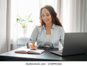 Adult Smiling Brunette Woman Doing Notes In Daily Book With Opened Laptop. She Study Something With Online Course At Home