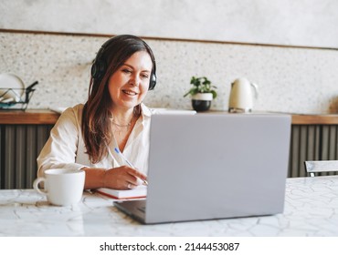 Adult smiling brunette woman fifty years in headphones doing notes in daily book with opened laptop,  study something with online course at home - Powered by Shutterstock