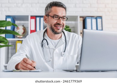 Adult smiling male doctor wearing glasses and white uniform with stethoscope working on laptop writing prescription doing notes sits at desk in his office workplace. Medical concept - Powered by Shutterstock