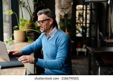 Adult smiling man in glasses and headphones typing on laptop while sitting in cafe