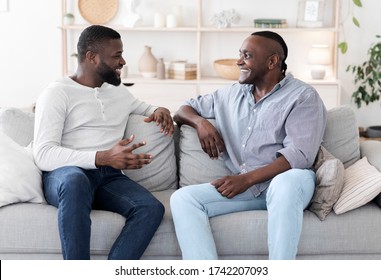 Adult Son And His Black Senior Father Talking While Relaxing On Couch At Home, Smiling To Each Other, Free Space