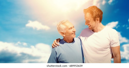 Adult Son Hugging His Old Father Against Cloudy Sky With Sunshine 