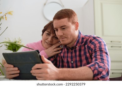 adult son patiently guiding his mother through the ins and outs of using a tablet. Tech Tutoring: Adult Son Helps Mother Master Tablet Skills - Powered by Shutterstock