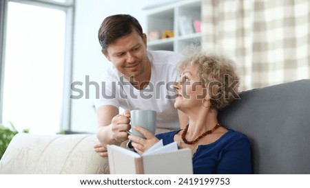 Adult son serves cup tea to an elderly mother. Relatives help each other during self-isolation. Time for reading fiction and books on self-development. Son pays attention to an elderly mother