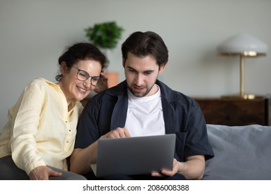 Adult son teaching mature mom to use online virtual app, banking service on laptop. Senior mother and grown child sitting on couch together, holding computer, browsing internet, talking