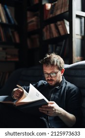 Adult Student Reading A Book In The College Library. Young Positive Male Wearing Glasses With Bookshelves On Background.