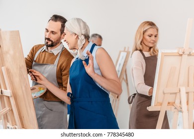 Adult Students In Aprons Painting On Easels During Art Class 