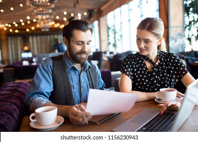 Adult stylish man and woman with papers and laptop sitting at table in cafeteria having cups of tea