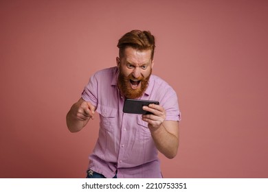 Adult Stylish Redhead Bearded Enthusiastic Man Shouting And Holding Phone Celebrating Victory, While Standing Over Isolated Coral Background