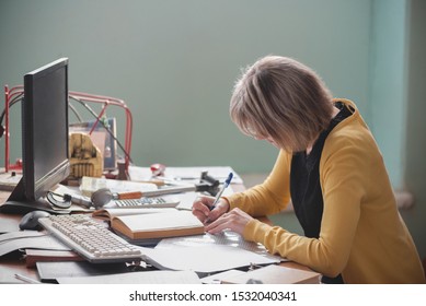 Adult Teacher Of College Technical Discipline Sitting By Her Desk And Is Writing A Class Book.