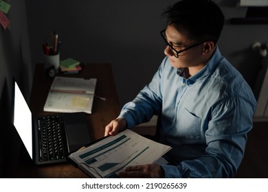 Adult Tense Asian Man With Documents And Laptop Working In Dark Room