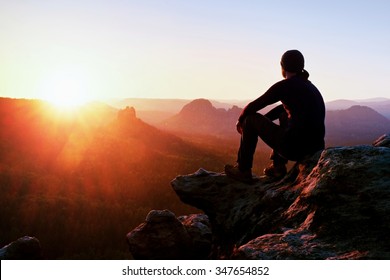 Adult tourist in black trousers, jacket and dark cap sit on cliff's edge and looking to misty hilly valley bellow - Shutterstock ID 347654852