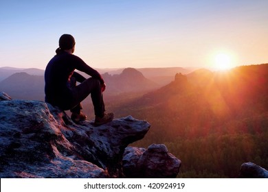 Adult tourist in black trousers, jacket and dark cap sit on cliff's edge and looking to misty hilly valley bellow - Powered by Shutterstock