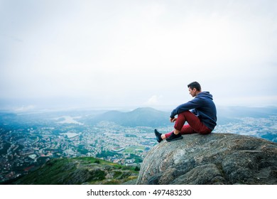 Adult tourist in red trousers, jacket sit on cliff's edge and looking to misty hilly valley bellow. Hiker relax and thinking alone in nature.