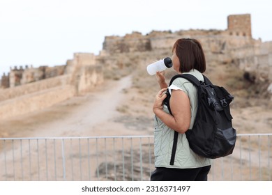 Adult traveling woman tourist with backpack drinks water while relax after walks, posing outdoors in ancient Europe fortress ruins. Retired people summer holiday vacation, active lifestyle concept. - Shutterstock ID 2316371497