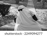 An adult using a white bedsheet as a cute ghost costume holds up peace signs with their hands while "haunting" a graveyard in black and white